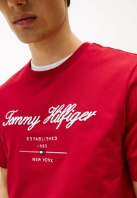Rød bomulds t-shirt med hvid broderet "Tommy Hilfiger" logo over "EETABLISHED 1985 NEW YORK," med rund hals og korte ærmer.