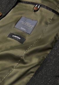 Das Innenfutter der schwarzen Jacke verfügt über ein olivgrünes Futter mit zwei Etiketten: "DIGEL" in Gold und "STRETCH" in Schwarz, sowie einem runden braunen Knopf.