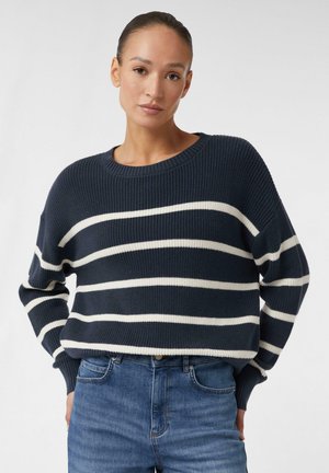 Marineblaues Strickpullover mit dicken weißen horizontalen Streifen, Rundhalsausschnitt und gerippten Ärmelabschlüssen. Getragen mit hellblauen Jeans.