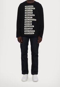 Maglietta nera a maniche lunghe con frasi bianche disposte a strati, abbinata a jeans in denim scuro e sneaker bianche.