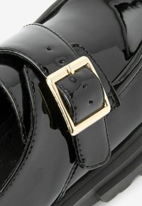 Scarpa in pelle verniciata nera con una larga fascia e una fibbia quadrata dorata, superficie liscia e suola in gomma texturizzata.