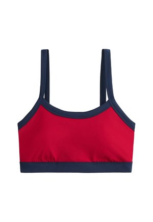 Giovane donna in piedi con pantaloni della tuta grigi, reggiseno sportivo rosso, sneakers beige, con le mani nelle tasche su uno sfondo grigio uniforme.