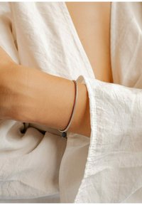 Pulsera de plata con un acabado suave, forma circular y diseño flexible, llevada en una muñeca al descubierto, contrastando con una tela blanca.