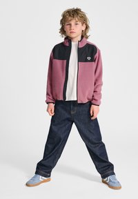 Fleecevest in roze en zwart met een hoge kraag, gecombineerd met donkerblauwe spijkerbroek en lichtblauwe sneakers met witte accenten.