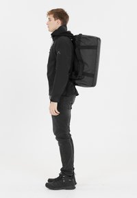 Musta reppu suorakaiteen muotoisella suunnittelulla, valmistettu kestävästä materiaalista, jossa on hihnat ja rakenteellinen muoto. Mallilla on päällään musta takki ja housut.