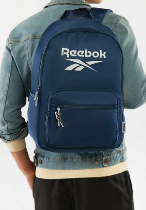 Persona que lleva una mochila azul de Reebok con un gran bolsillo frontal y logo blanco, combinada con una chaqueta de mezclilla clara y pantalones negros.