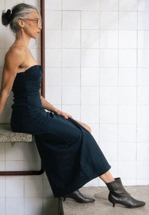 Navyblauwe strapless maxi-jurk met gestructureerde stof, zijsplit en ruches, gecombineerd met bruine leren enkellaarsjes op een grijze ondergrond.