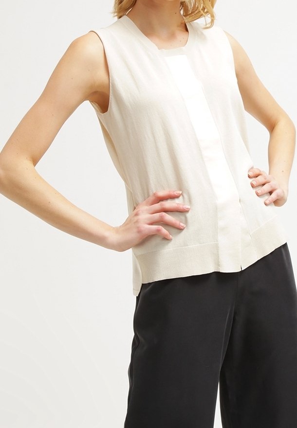 Ärmlös beige topp med texturerad stickning, med en central vertikal panel och ribbad fåll, i kombination med svarta byxor.