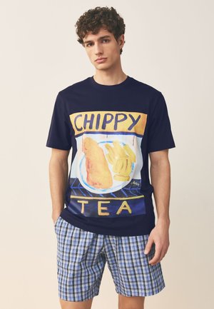 Młody mężczyzna w granatowej koszulce z grafiką ryby i frytek oraz tekstem "CHIPPY TEA", w połączeniu z niebieskimi, kratowanymi szortami.