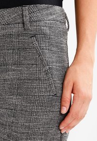 Pantalon gris tissé avec un motif texturé, doté d'une poche latérale et d'une coupe ajustée. Le tissu semble structuré avec une légère brillance.