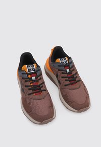 Baskets brunes et grises avec des matériaux en mesh et en suede, lacets noirs et rouges, accents orange et un logo sur la languette.