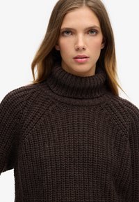 Brązowy sweter z wysokim kołnierzem, dziany w splot w teksturę z prążkami i rękawami raglanowymi, o luźnym kroju.