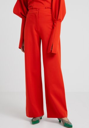Femme portant un pantalon large taille haute rouge vif associé à un haut manches longues assorti et des chaussures vertes à motifs à petit talon.