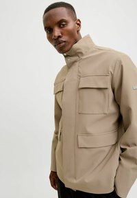 Bežova, vodoodporna jakna z visokim ovratnikom, dvema sprednjima žepoma in gladko teksturo. Ima zapiranje na klešče in sodoben dizajn.