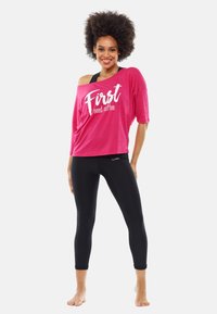 Leende kvinna med afro som bär en rosa off-shoulder-tröja med texten "Först behöver jag kaffe" och svarta leggings, stående barfota.