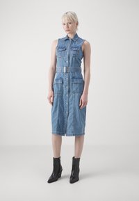 Abito midi in denim con design senza maniche, colletto, bottoni frontali e due tasche sul petto. Presenta una vita con cintura e orlo sfilacciato. Stivali neri con tacco.