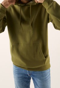 Felpa verde con cappuccio, dotata di tasca frontale a marsupio e cordino, realizzata in un morbido tessuto misto cotone. Indossata sopra a jeans in denim blu.