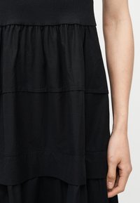 Κοντινή λήψη μαύρου φόρεματος με στρώσεις, με λεπτομέρειες μαζεμένου υφάσματος και ορατό χέρι που ακουμπά δίπλα στο σώμα.