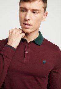 Polo bordeaux à manches longues avec col vert, doté d'une patte de boutonnage à trois boutons et d'un petit logo vert sur la poitrine. Finition contrastante sur les manches.