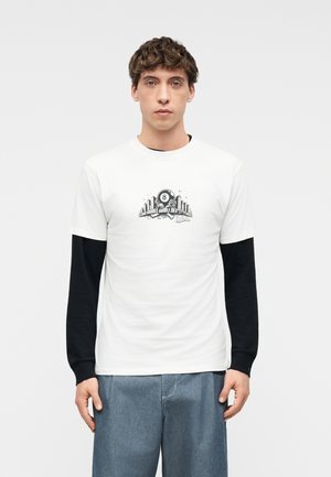 Mladý muž s kučeravými vlasmi, oblečený v bielom tričku cez čierne dlhé rukávy, na tričku je čierno-biela grafika s osmičkou a mestskou scenériou, modré džínsy.