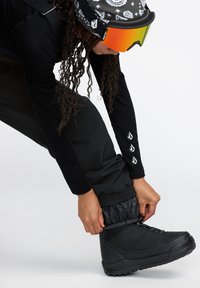 Zwarte sneeuwlaarzen van textuurmateriaal, met accentlogo en een veilige sluiting. Draagt zwarte kleding met decoratieve armdetails.