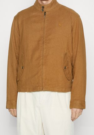 Braune Jacke mit Reißverschluss, knöpfbarem Kragen und Klapptaschen, getragen über weiße Hose, Teilansicht einer Person vom Hals bis zu den Hüften.