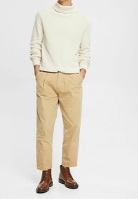 Hellbeiger Rollkragenpullover mit strukturierter Strickoptik, kombiniert mit hellbraunen, schmal zulaufenden Chinos und braunen Schnürstiefeln bis zum Knöchel.