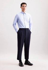 Lichtblauw patroonhemd met kraag, gecombineerd met marineblauwe nette broek en zwarte leren schoenen. Model staat met handen in de zakken.