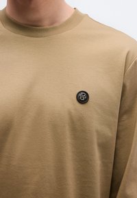 Bézs színű rövid ujjú póló kerek nyakkivágással és fekete kör alakú logóval a mellrészen, sima, könnyű anyagból.