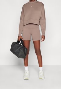 Persona che indossa pantaloni sportivi e felpa marroni abbinati, scarpe da ginnastica bianche e tiene una borsa da palestra nera.