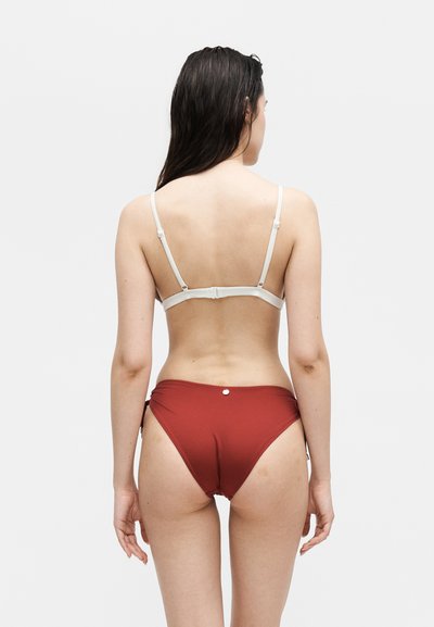 Γυναίκα που φοράει λευκό τοπ μπικίνι και μαγιό με ψηλόμεσο κατώτατο μέρος σε χρώμα σκουριάς, απεικονίζεται από πίσω σε απλό ανοιχτό φόντο.