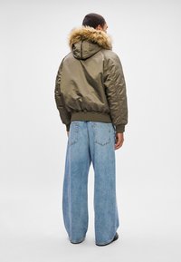 Hombre vestido con una chaqueta acolchada verde oliva con capucha de piel y pantalones vaqueros de pierna ancha de color azul claro, de pie de espaldas frente a un fondo blanco liso.