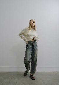 Suéter recortado beige, de corte relajado, combinado con jeans de mezclilla azul claro de cintura alta y mocasines marrón oscuro, sobre un fondo gris liso.