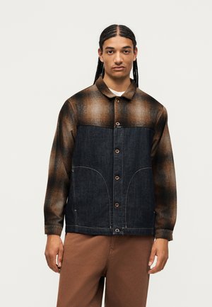 Man met gevlochten haar die een bruin-zwart geruite jas draagt met een denim paneel en een bruine broek, staand tegen een effen achtergrond.
