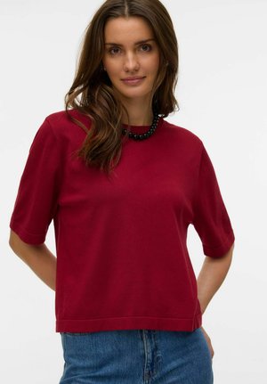 Kvinde med langt brunt hår iført en rød kortærmet top, blå jeans og en sort perlehalskæde, stående med hænderne bag ryggen.