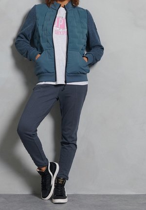 Person iført en blå quiltet jakke, grå joggingbukser og sorte sneakers, der står op ad en ensfarvet grå væg.