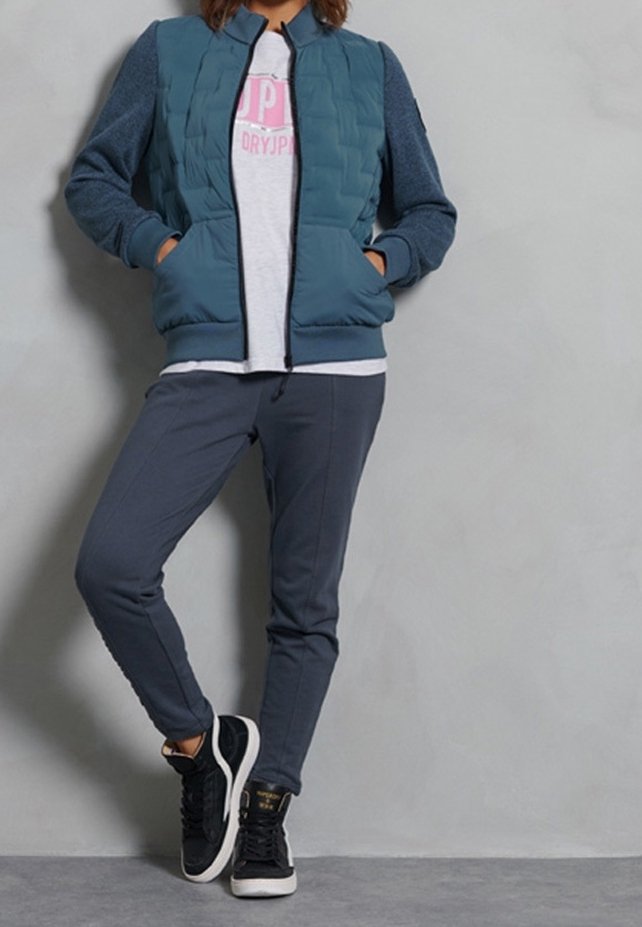 Personne portant une veste matelassée bleue, un pantalon de survêtement gris et des baskets noires, debout contre un mur gris uni.