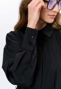 Schwarzes Hemd mit Knöpfen, einem Kragen, gepufften Ärmeln und zwei schwarzen Knöpfen an den Manschetten. Glattes Gewebe mit subtiler Glanz.