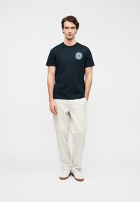 Μπλε βαμβακερό μπλουζάκι με στρογγυλή λαιμόκοψη και κοντά μανίκια, με ένα γαλάζιο κυκλικό γραφικό στο αριστερό στήθος. Συνδυάζεται με μπεζ παντελόνια.