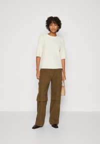 Cremfarbener, gerippter Strickpullover mit kurzen Ärmeln, kombiniert mit braunen Cargo-Hosen mit einem geschichteten Design. Das Model hält eine kleine beige Handtasche.