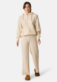 Beżowy, prążkowany hoodie z kieszenią z przodu i sznurkiem, w połączeniu z dopasowanymi, prążkowanymi spodniami o szerokim kroju i brązowym obuwiem wsuwanym.