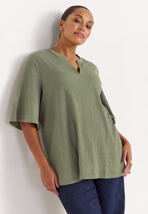 Kobieta w luźnej oliwkowozielonej bluzce o fakturze, z rękawami do łokcia, i ciemnoniebieskich spodniach, stojąca na jasnym tle.