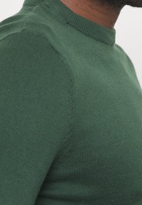 Mörkgrön stickad tröja med rund halsringning, ribbat mönster och figursydd design. Tyget verkar mjukt och elastiskt.