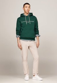 Sudadera verde con bolsillo delantero, logo blanco y cordones. Llevada sobre una camisa azul a cuadros. Combinada con pantalones beige claros y zapatillas blancas.