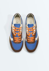 Paire de baskets bleues, blanches et marron avec des lacets orange vif, vues de dessus sur un fond clair uni.