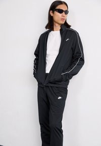 Une personne se tient devant un fond uni, portant un survêtement noir de Nike et des lunettes de soleil, les mains dans les poches et une expression confiante.
