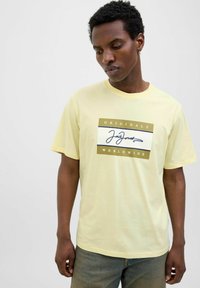 Lys gul kortærmet t-shirt med et centralt grafisk motiv, der viser "ORIGINALS" og "WORLDWIDE" i guld og mørkeblå, med en glat bomuldsstruktur.