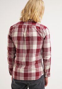 Personne blonde portant une chemise à manches longues à grands carreaux rouges et blancs, avec un pantalon gris foncé, vue de dos.