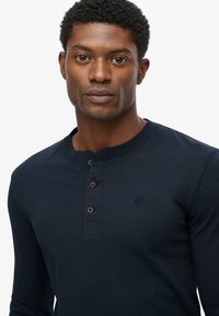 Marinefarbener Langarm-Henley-Shirt aus weichem Stoff, mit einem Knopfverschluss aus braunen Knöpfen und einem kleinen gestickten Logo auf der Brust.
