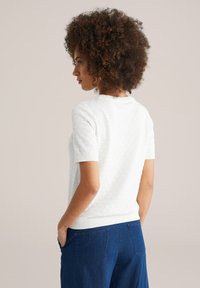 Kvinna med lockigt hår som bär en vit texturerad kortärmad topp och blå jeans, stående med händerna i fickorna, vänd åt vänster.